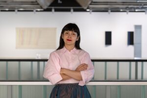 A brown woman with long dark hair and bangs stands with arms crossed, wearing a light pink shirt and dark skirt. The background is a modern, minimalistic space with abstract art on the walls, suggesting an art gallery setting. The person has a calm expression and red lipstick.