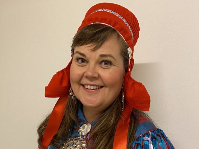 Woman wearing a Sámi national dress and a red headdress and smiling at the camera.