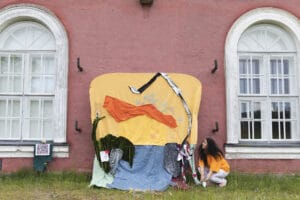 A woman with black curly hair looking up at a playful fabric artwork outdoors next to the HIAP Suomenlinna building with pink walls and white windows. Taken in the springtime.