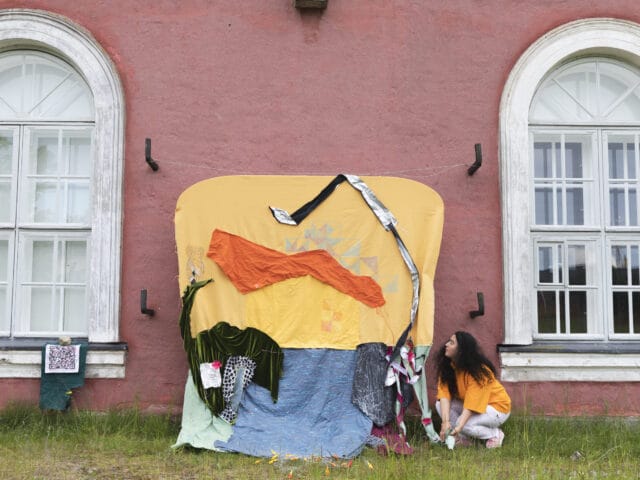 A woman with black curly hair looking up at a playful fabric artwork outdoors next to the HIAP Suomenlinna building with pink walls and white windows. Taken in the springtime.