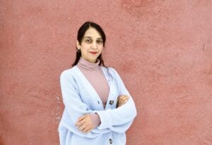 Brown woman with long hair and crossed arms standing against a pink wall. She is wearing a pink turtleneck and a light blue sweater. She is smiling.